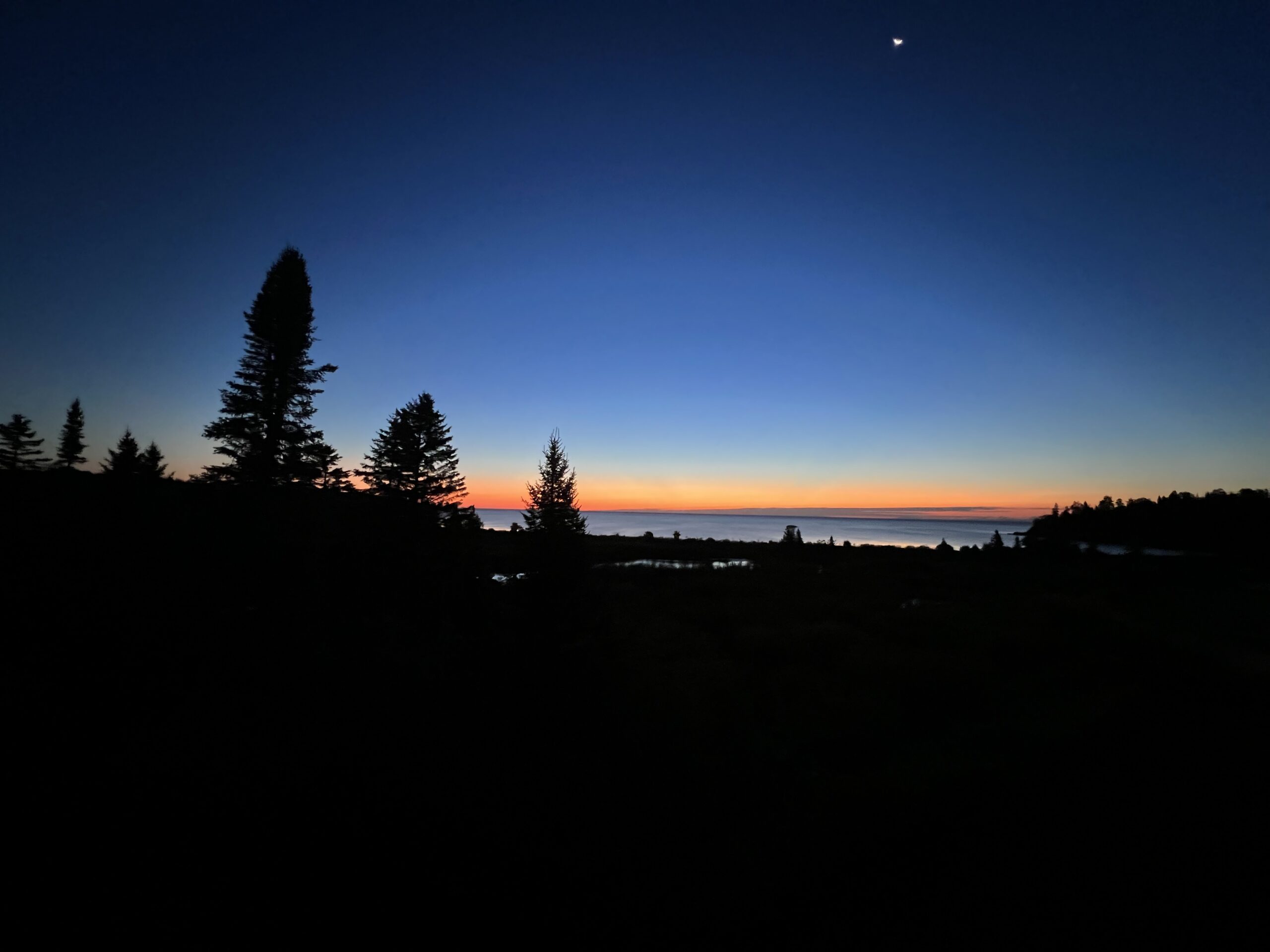 Dawn over Lake Superior with a dark evergreen forest in the foreground, soft pink and peach hues rising at the horizon, and deep blue water reflecting the first light—symbolizing clarity, reflection, and new beginnings for The Sober Globetrotter.