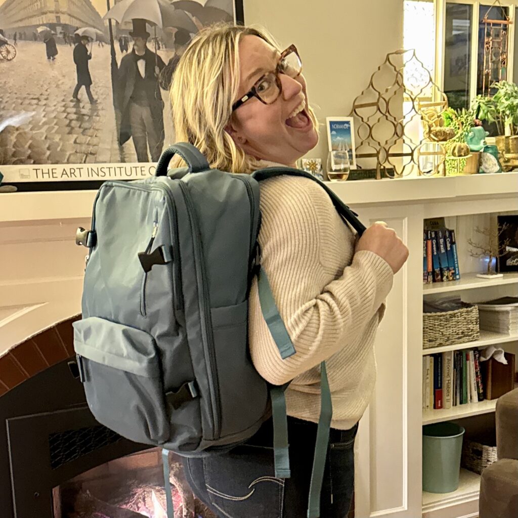 Ginny stands in her living room wearing her perfect travel backpack in grey-teal, looking over her shoulder at the camera with a big smile.
