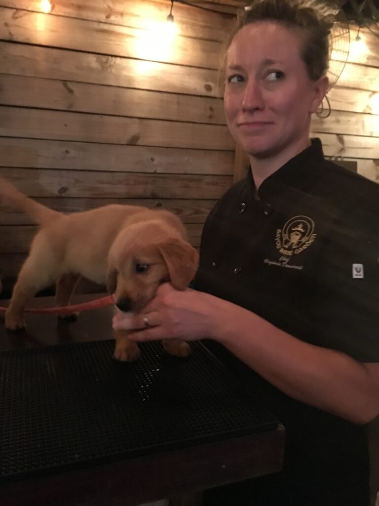 Ginny Dee in a short-sleeve chef coat, next to a puppy standing on the bar with her hand in its mouth—early days in the kitchen, full of curiosity and chaos.