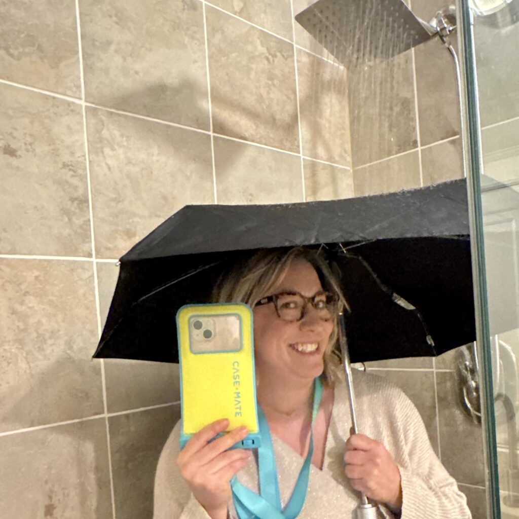 A smiling Ginny holds up her phone in its bright yellow case with blue accents and neck strap, as she stands under an umbrella in the shower (which is on).