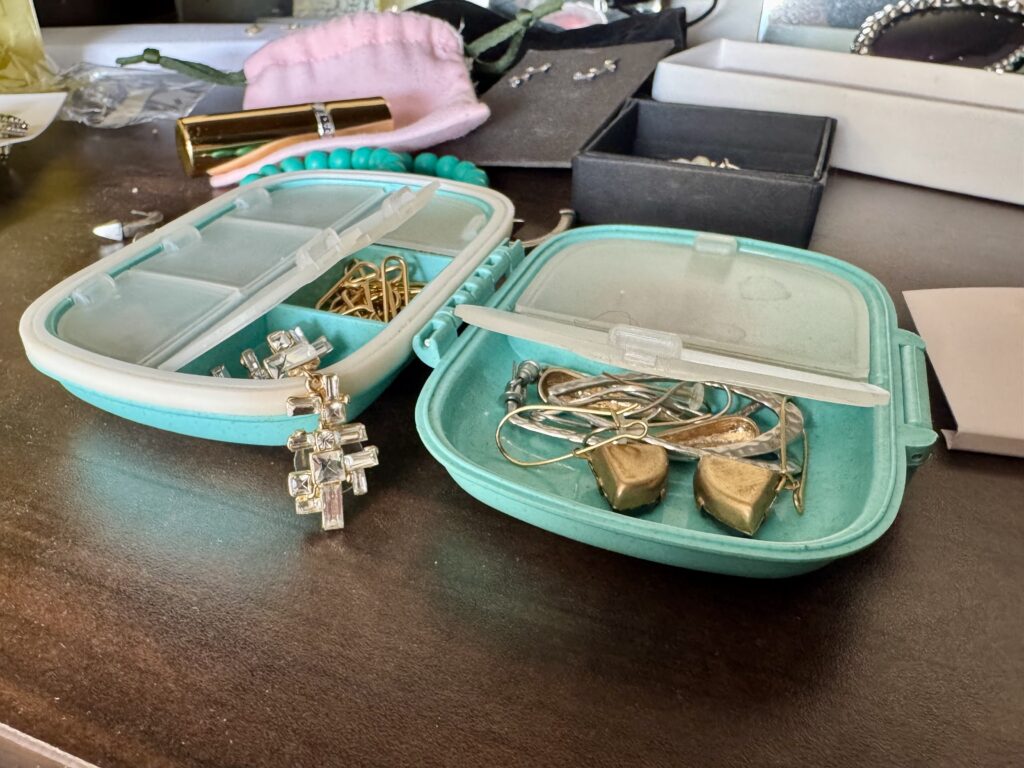 A teal pill box with frosted-clear dividers spills out various jewelry onto a wooden dresser top.