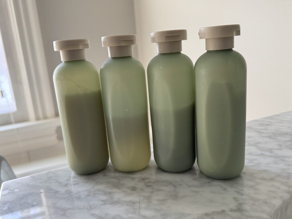 Four large, green toiletry bottles with tan caps sit on a marble countertop. We can see that they are all various levels of full.