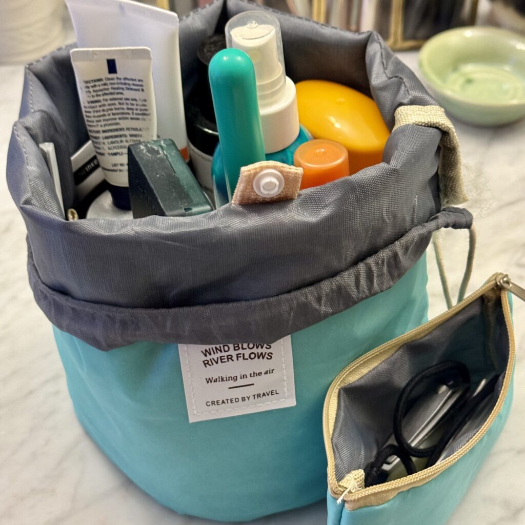 Teal bucket back with the top folded down to reveal the tops of various toiletries, with a small zippered pouch open beside it.