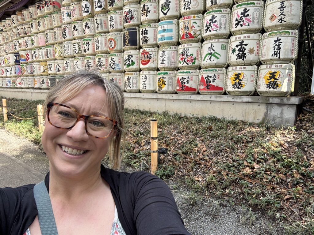 Ginny Dee smiling in front of a wall of vibrant, decorative sake barrels in Tokyo—joyful and grounded, embracing sobriety and wonder.