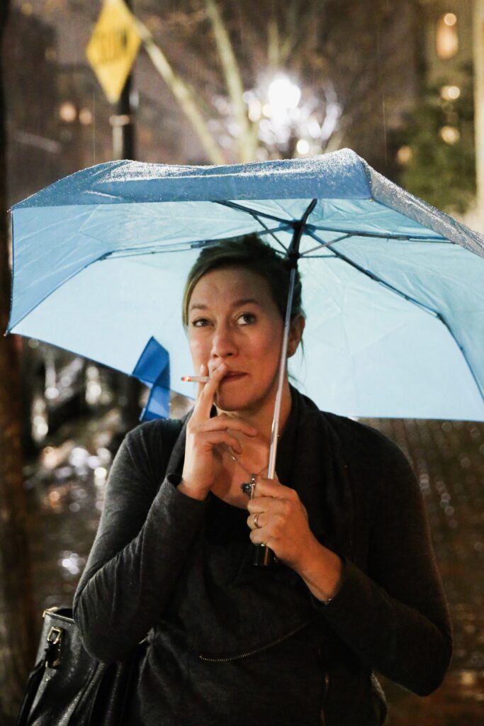 Ginny Dee under a bright blue umbrella, smoking a cigarette on a rainy night, with glistening wet pavement and soft city lights in the background.