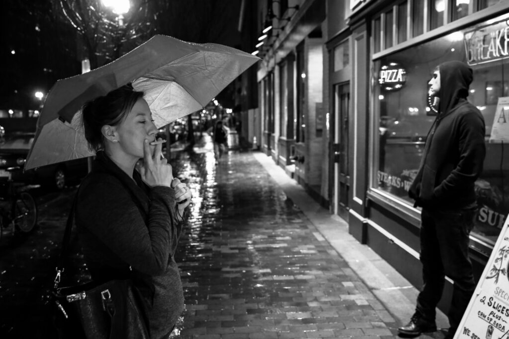 Black-and-white photo of Ginny Dee standing under an umbrella, smoking outside a restaurant after rain—capturing the gritty exhaustion of her old life.