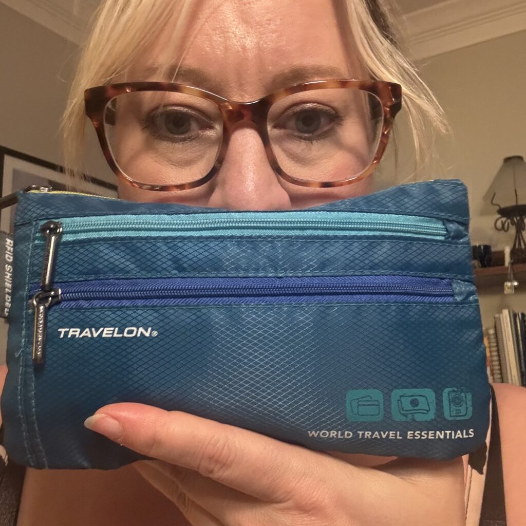Ginny holds up a blue pouch with two front zippers and one (barely visible) on top. The bag is positioned in front of her mouth, with eyes and nose visible above.