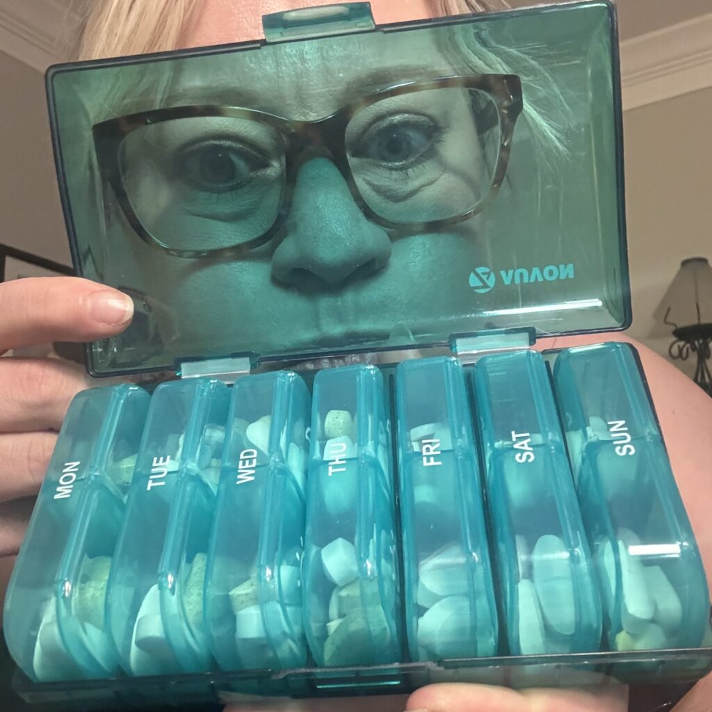Ginny making a funny face behind the clear-turquoise open top of the large pill box, showing the 7 2-compartment mini cases full of pills.