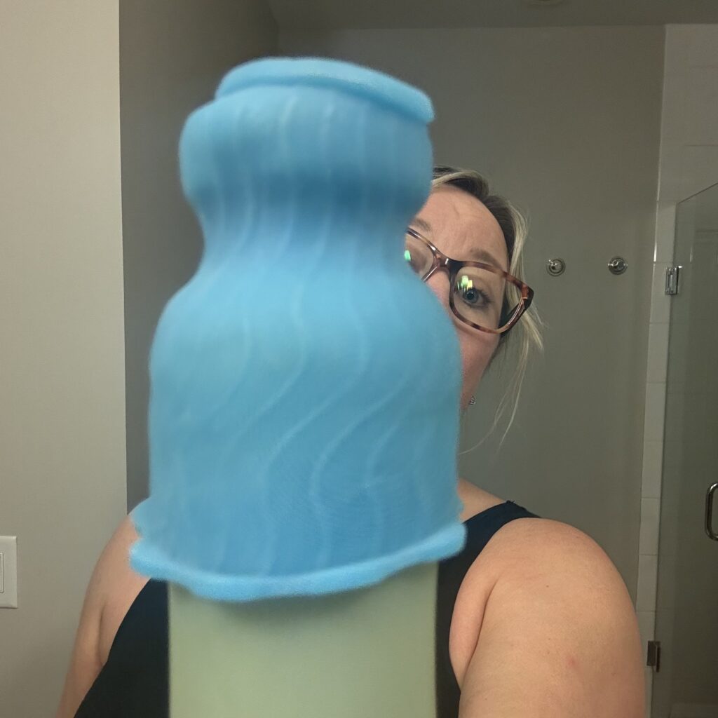 Ginny holds out a green toiletry bottle, the top covered with the blue silicone sleeve that will keep it from leaking, as she peers around the edge from the background.