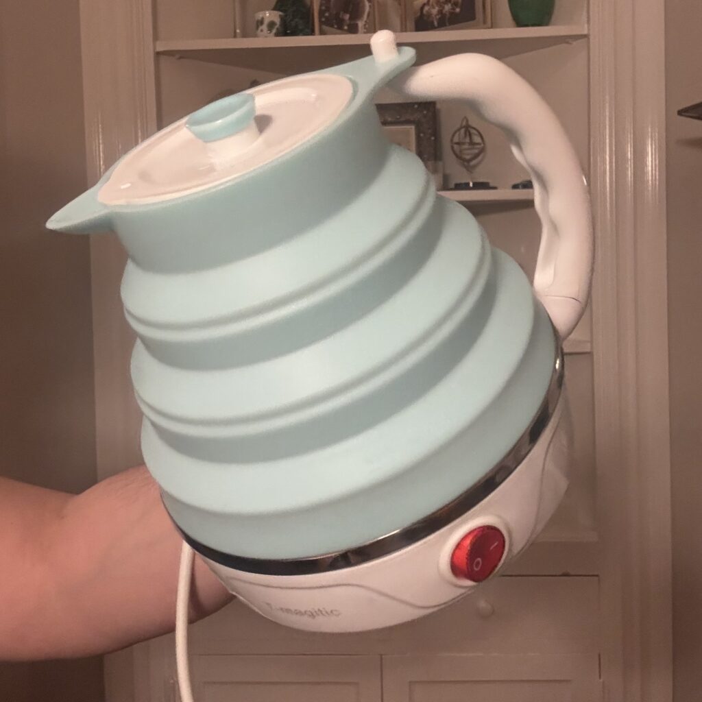 Ginny holds up the expanded light-blue electric kettle, white plastic handle rotated up and hooked through the top of the silicone sides to give it stability.