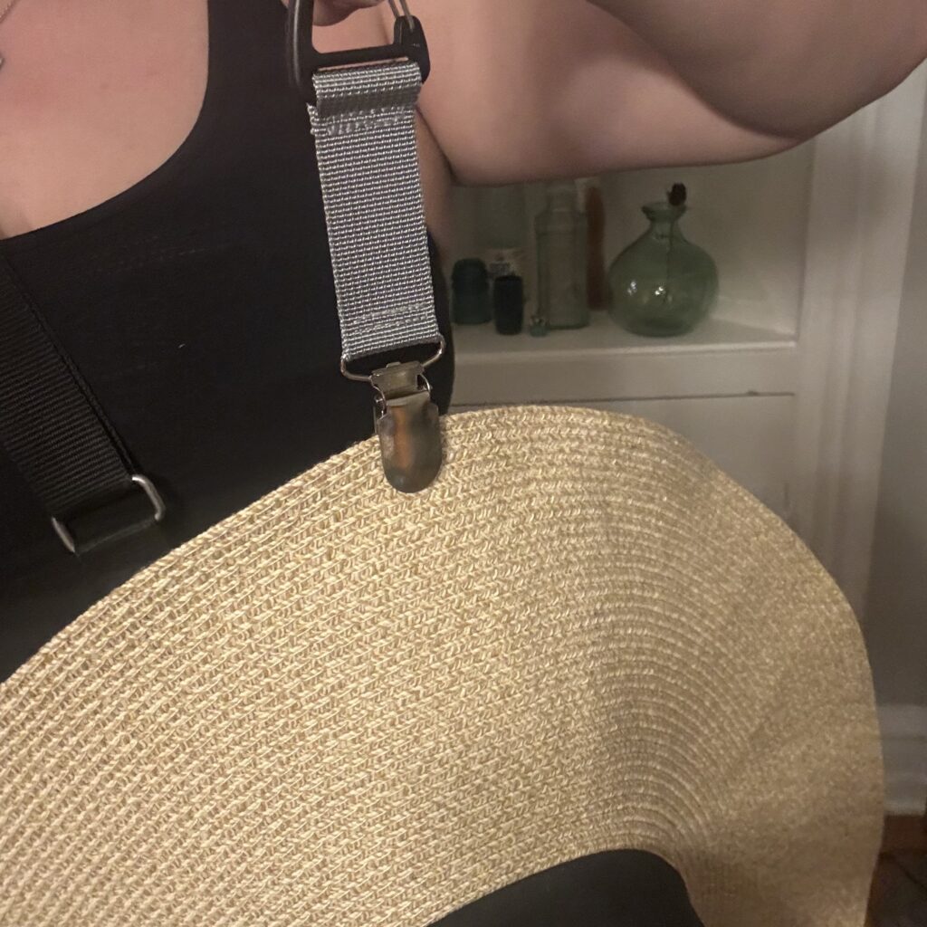 Ginny holds up one end of the hat clip (which clips easily around any bag strap), while the other holds up a large tan straw hat.