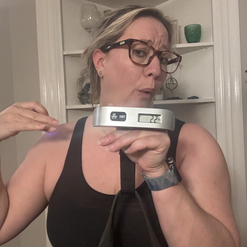 Ginny makes a weird face as she holds up the luggage scale, supporting a bag which we can see weighs 2.2 lbs.