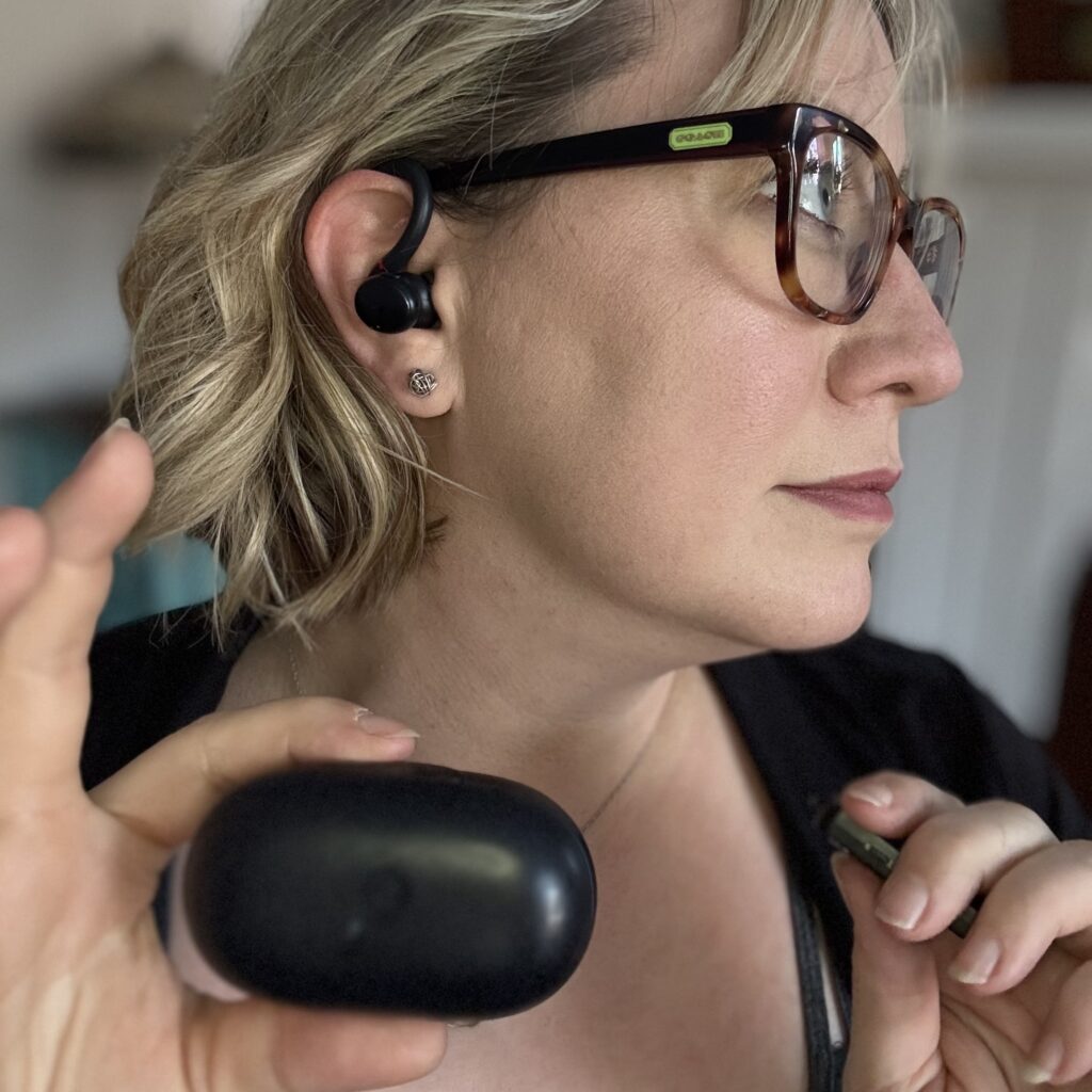 Profile view of Ginny wearing the ear buds with over-ear hook and holding up their charging case.