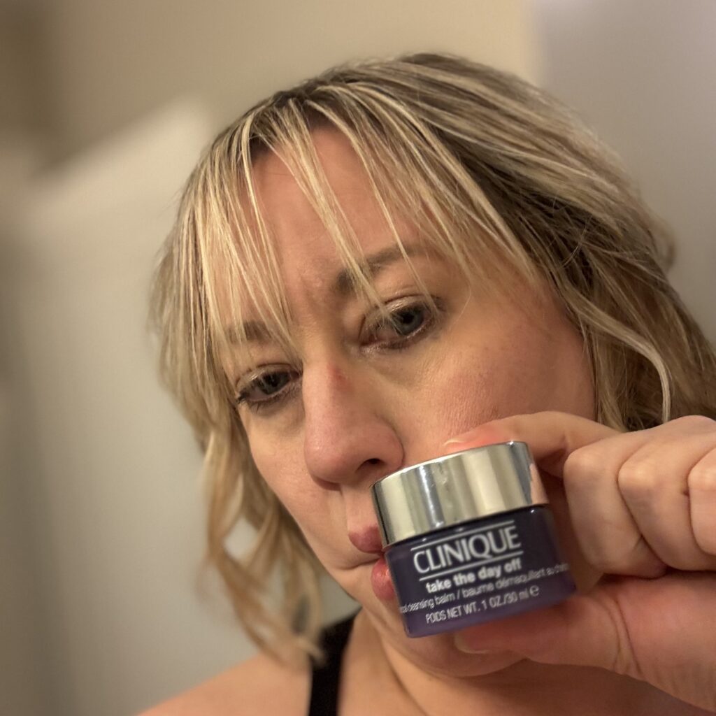 Ginny at the end of a long day, looking tired, with slightly smudgy makeup, holding up the small purple tub of Clinique Take The Day Off balm