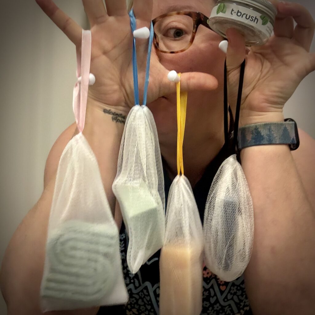 Ginny Dee holding up eco-friendly travel toiletries in mesh bags—solid shampoo, conditioner, soap, and toothpaste tabs—perfect lightweight alternatives for packing light.