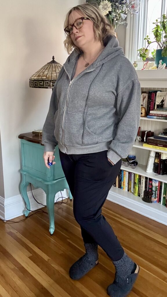 Ginny ready for a nap in a thick, oversized grey sweatshirt, black joggers, fuzzy dark grey socks, and fuzzy dark grey clogs