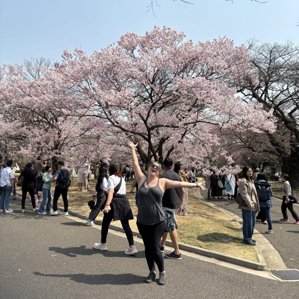 Ginny smiling with arms raised under a blooming cherry tree in Japan—embracing joy in the moment, crowds and all
