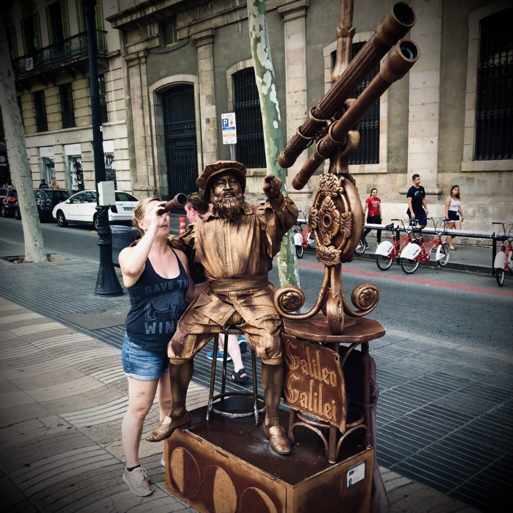 On Las Ramblas in Barcelona, Ginny Dee stands beside a bronze street performer dressed as Galileo. Holding a handheld telescope, she gazes playfully into the distance — as if scouting new travel destinations — while Galileo points the way, surrounded by props and moon phases.