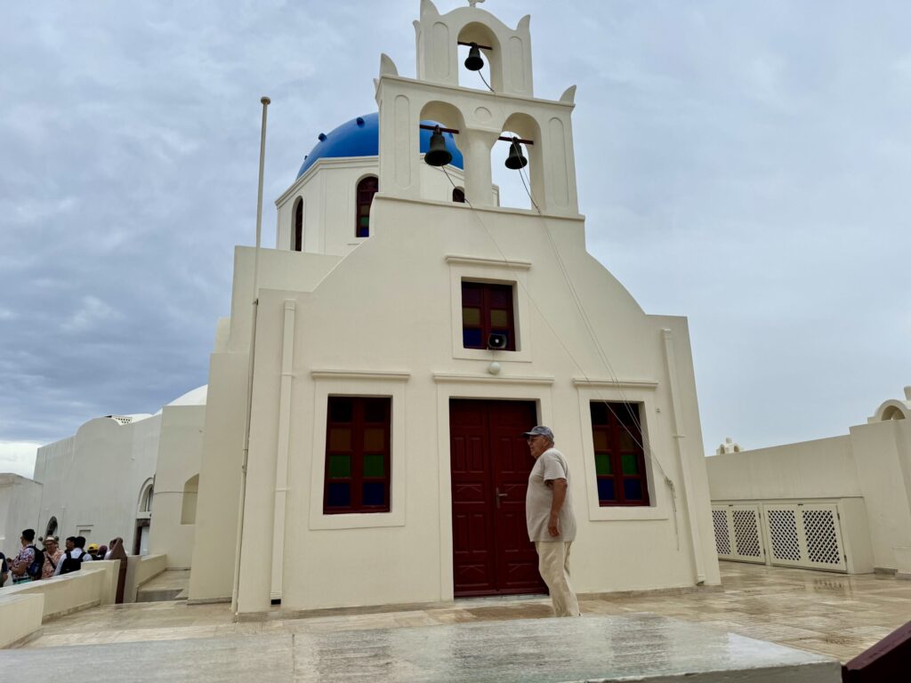 Front view of the Church of Panagia Platsani in Oia’s main square under cloudy skies, its white facade appearing tan in the dim light; a man in tan stands in front, blending almost perfectly with the rain-dimmed facade, beneath three bells and a blue dome.