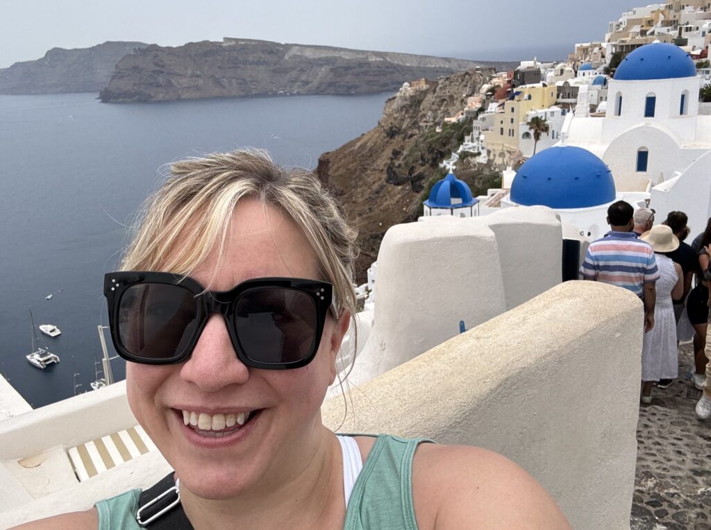Ginny smiling in sunglasses and a teal tank top in a selfie with the iconic three blue domes of Oia behind her; cliffs and the sea stretch out to the left, while a line of tourists waits for photos to the right.