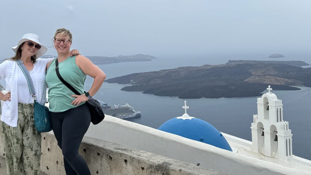 Ginny and travel companion L standing in front of a low wall overlooking Firostefani’s blue dome and three bells, with the Aegean Sea, a low island, and a grey sky behind them; Ginny wears a teal tank top with visible goosebumps, and L’s hair is windswept