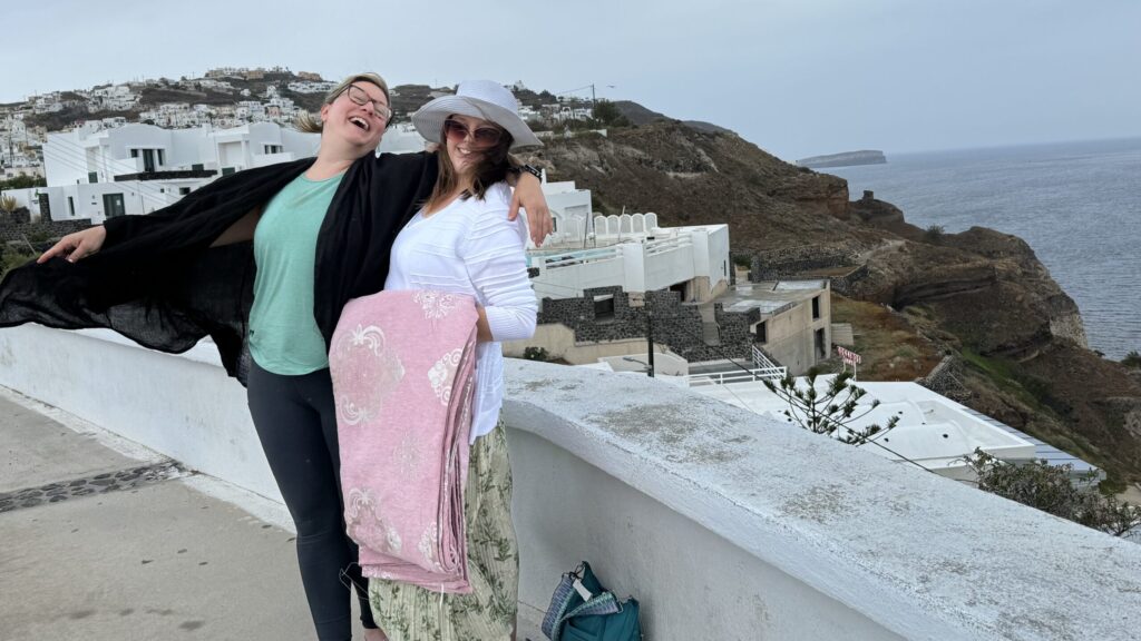Ginny and L at a scenic overlook after a textile shop stop; Ginny wears a black shawl caught in the wind like a wing, head flung back and arm out in a dramatic pose, while L holds a pink blanket and smiles; the cliffside and white village of Santorini rise behind them