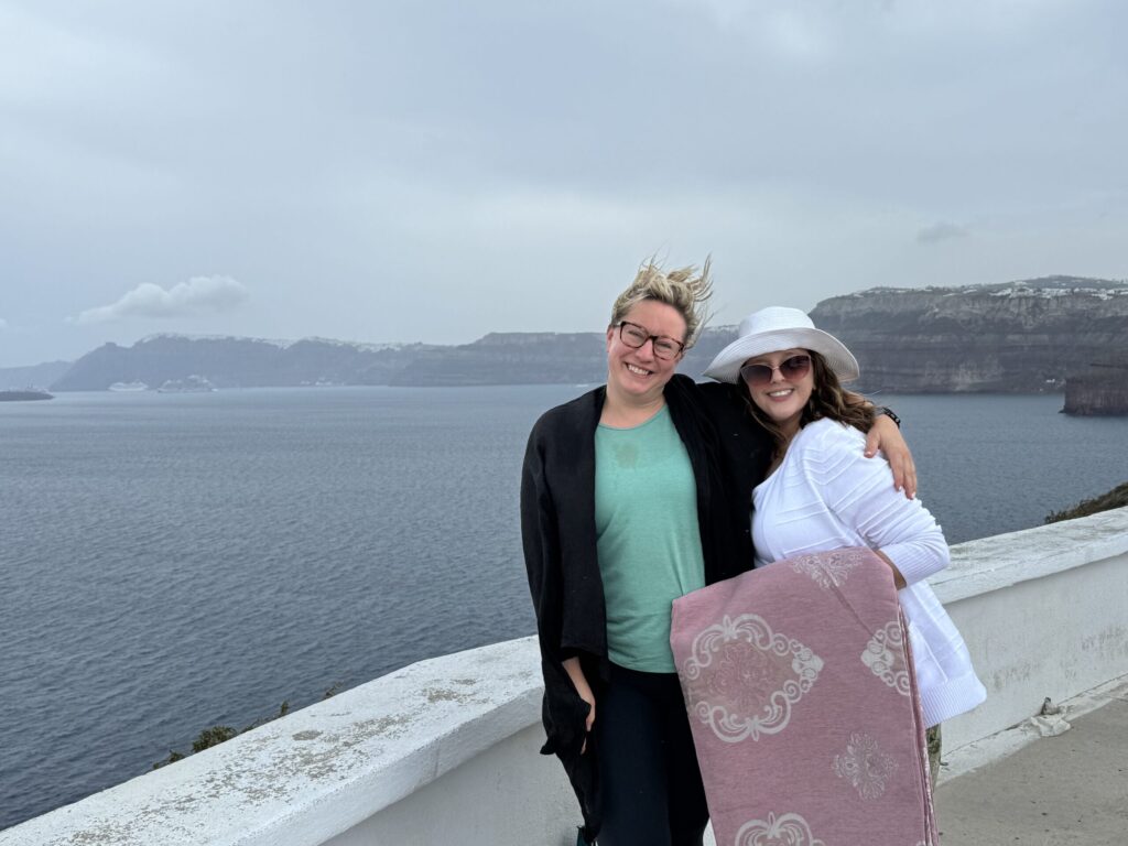 Ginny and L smiling at a viewpoint facing the opposite direction, with the full curve of Santorini’s caldera cliffs visible in the background; the wind is so strong it’s blowing Ginny’s hair straight up into the air