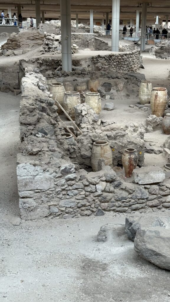 Wide view across the Akrotiri ruins showing low stone walls outlining multiple ancient buildings; several large reassembled storage jars (pithoi), some with traces of original paint, sit in one structure; modern support pillars for the roof are visible among the ruins in the background.