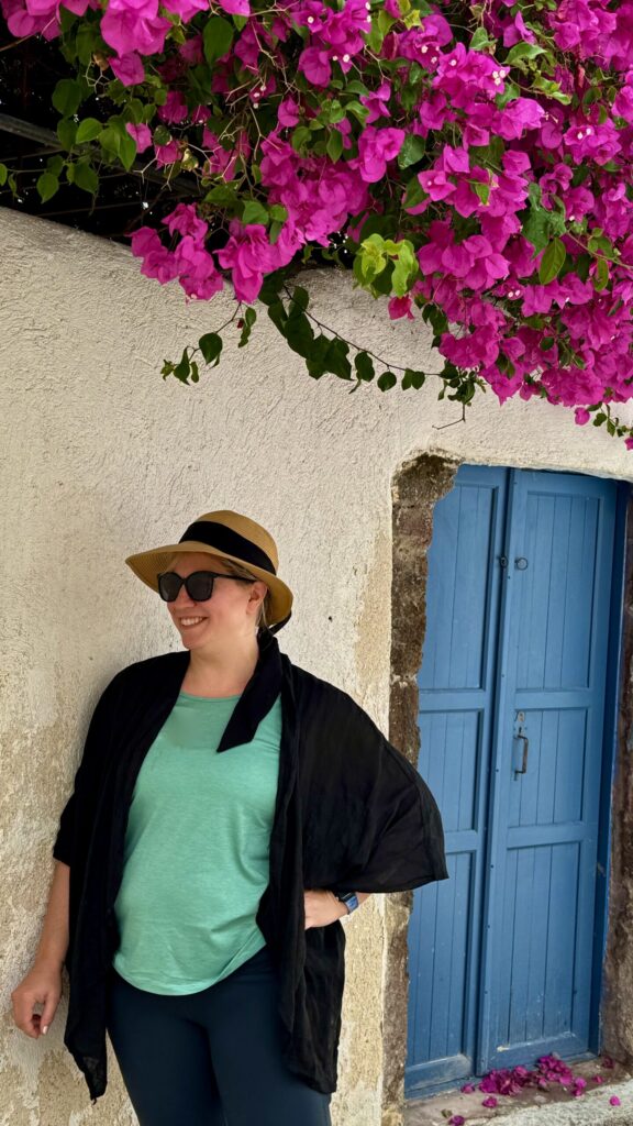 Ginny in sun hat and sunglasses standing beside a bright blue door and blooming pink bougainvillea in Emporeio, Santorini