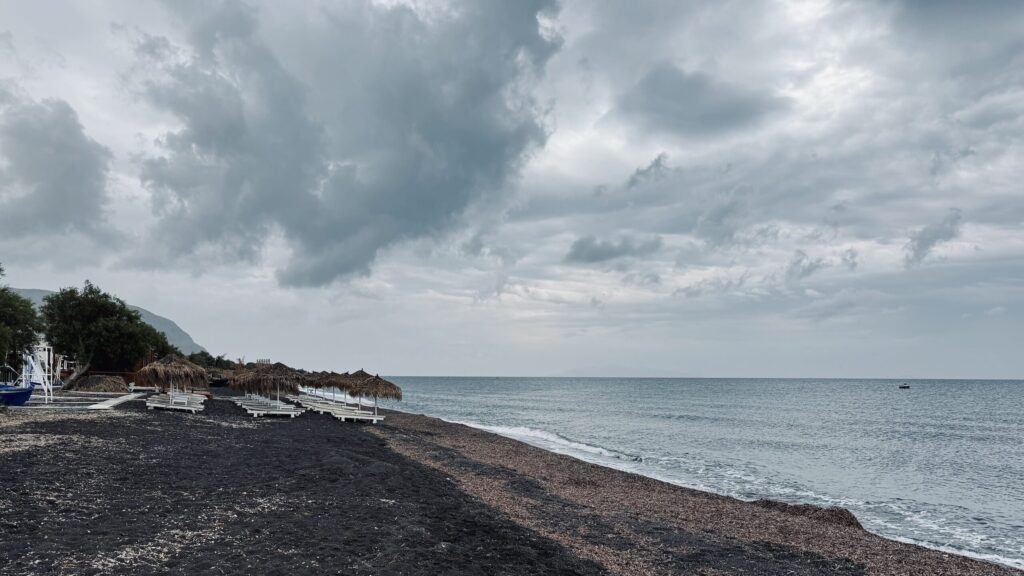 View of Perivolos Beach with a dramatic cloudy sky filling the top two-thirds of the image; the lower third shows silvery blue sea, a stripe of reddish shoreline debris, and black volcanic sand; to the left, rows of white beach chairs beneath palm umbrellas are visible near a few trees, with a distant shadowy cliff on the horizon