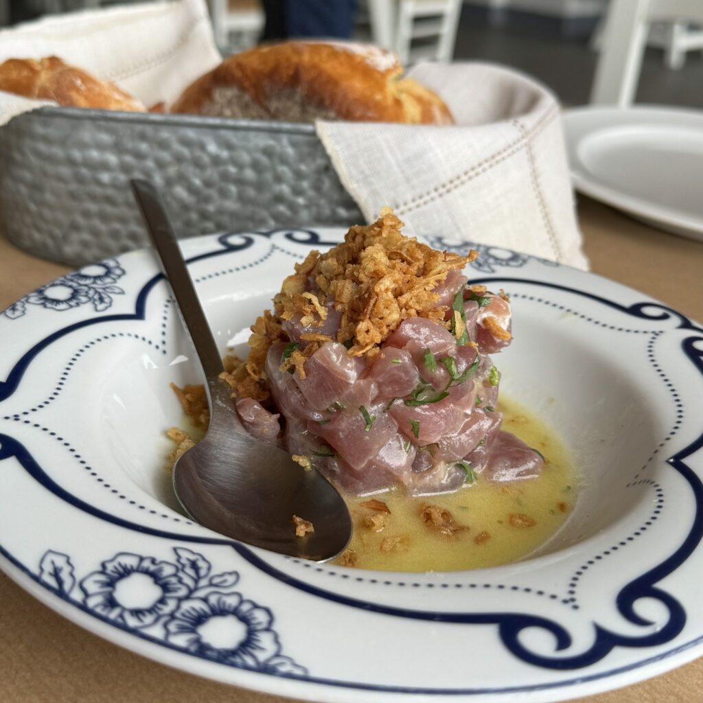 Tuna ceviche served in a cylindrical stack topped with crispy onions, sitting in a yellow citrus sauce in a shallow blue and white patterned bowl; basket of bread is visible in the background