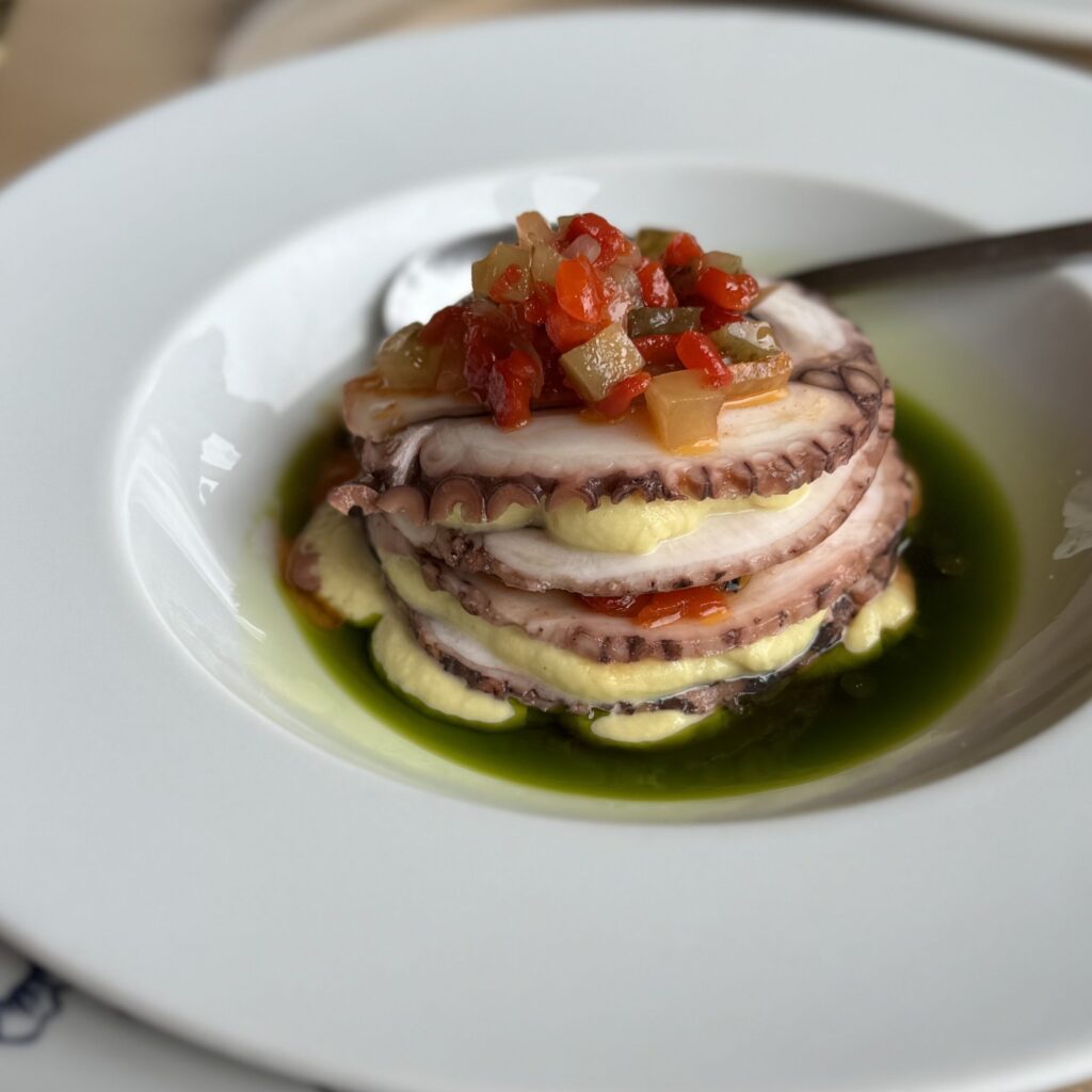 Tower of thinly sliced octopus with white flesh and purplish skin stacked with pale yellow-green fava puree, topped with finely diced red and yellow peppers; served in a shallow white bowl with herbed green oil pooling at the bottom