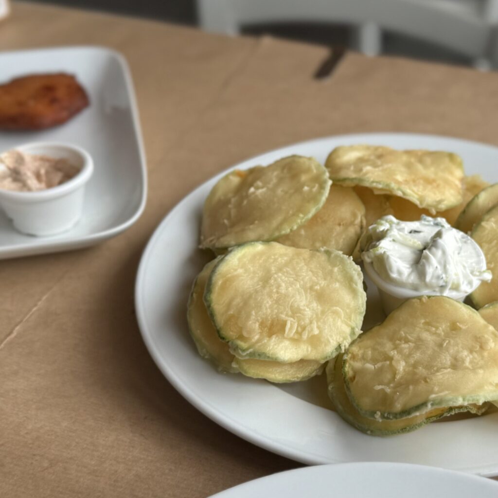 White plate of thin, golden-fried zucchini slices in a light, translucent batter with a tall dollop of thick white tzatziki in the center; in the background, a rectangular white plate holds a round tomato fritter and a small dish of pink aioli