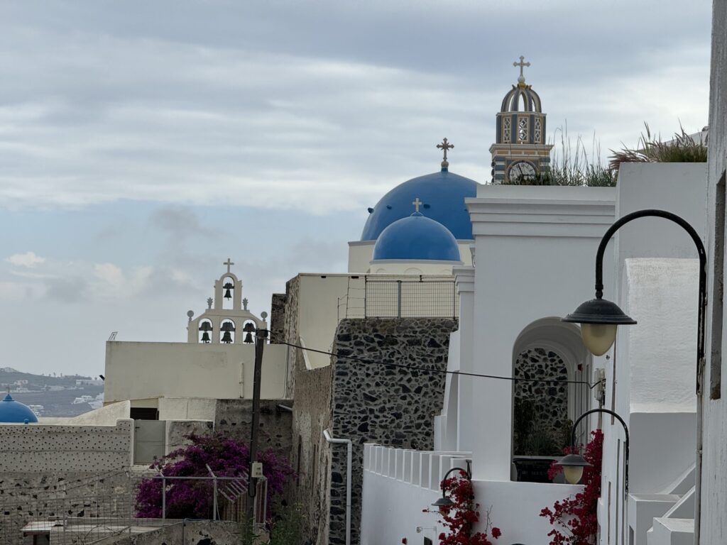 Layered view of Fira with a curved antique lamp on a white building in the foreground, decorated with red foliage and rooftop grass hinting at the steep slope; behind it, a dark stone wall, two blue domes, the Orthodox cathedral’s clock and bell tower, and a pale wall topped with three bells in arched niches; in the distance, another blue dome and cliffside buildings dot the dark landscape beneath a sky threatening to break into sunlight