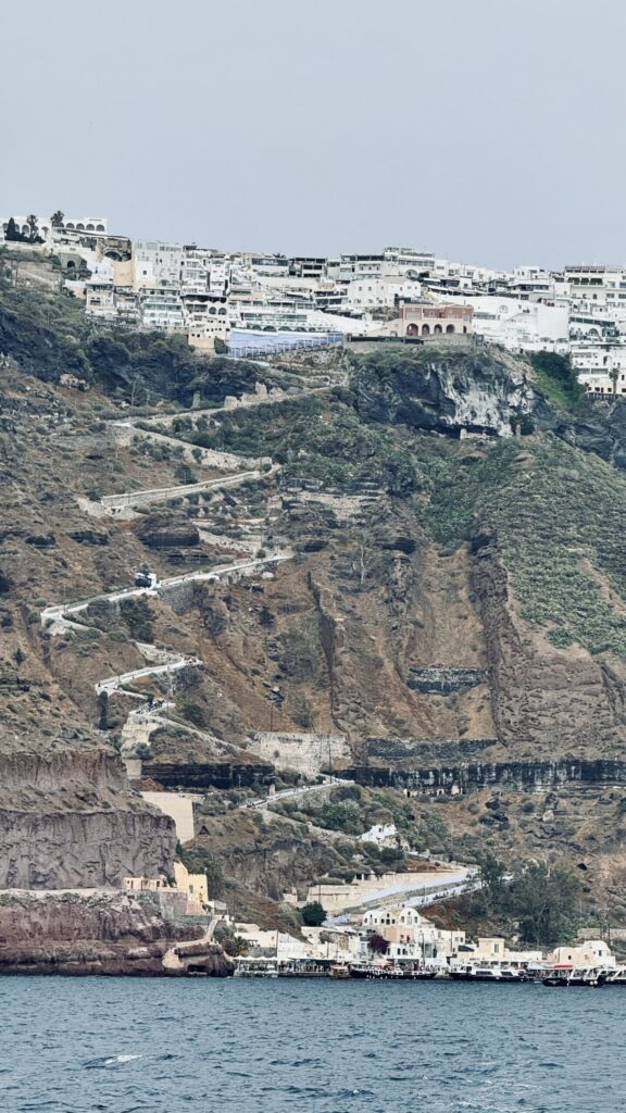 View from the tender boat of Santorini’s steep cliff face between Fira and the Old Port; at the top, the white buildings of Fira cluster along the rim, while a smaller whitewashed village sits at the base; between them, layered red and brown volcanic rock with black strata lines is dotted with green patches and crisscrossed by the winding Karavolades Stairs; a rippling blue sea fills the bottom of the image, with a band of white clouds above