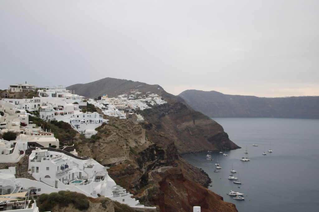 View of Santorini’s cliffside layered with red, brown, and black rock, topped by whitewashed buildings above a grey-blue sea dotted with sailboats, with a dim grey sky and a pink glow on the far caldera rim.