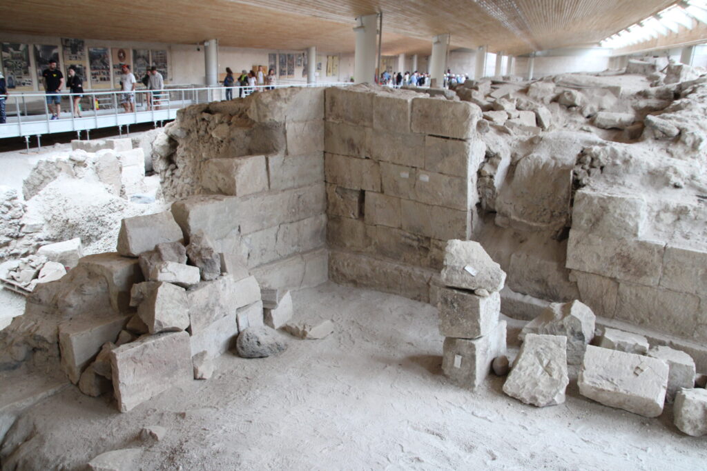 Inside the Akrotiri archaeological site, showing the corner of a light grey stone-block building in the foreground with visible notches and interlocking joints designed for earthquake resistance; rubble and a raised walkway with tourists are visible in the background beneath the modern building’s interior walls.