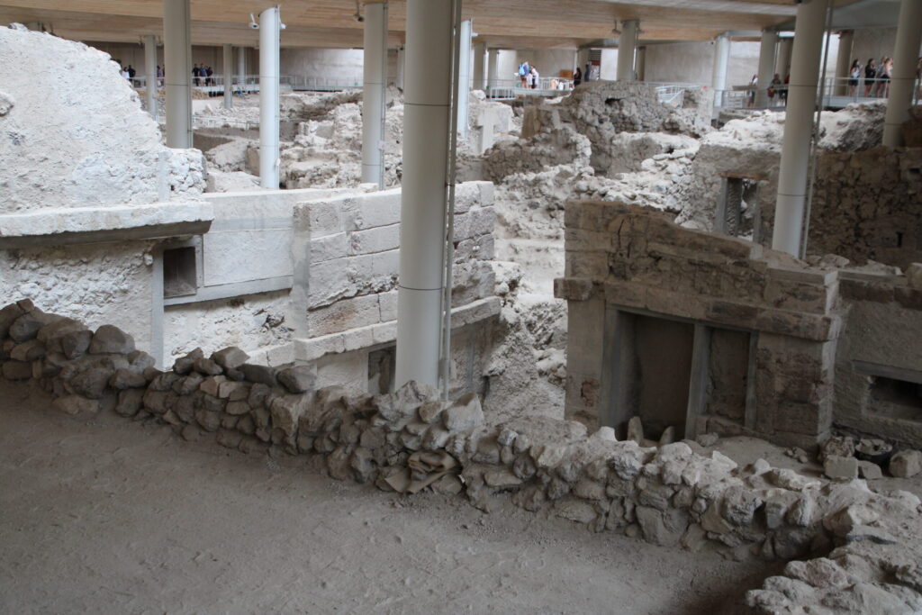 Wide view of Akrotiri ruins highlighting several preserved multi-story structures; at right, a two-story wall with a wooden door frame and window openings, and at left, a rounded tower with smooth stone blocks rising to a domed top; in the foreground, remnants of a second-floor room bordered by low walls, with more ruins and modern roof supports in the background