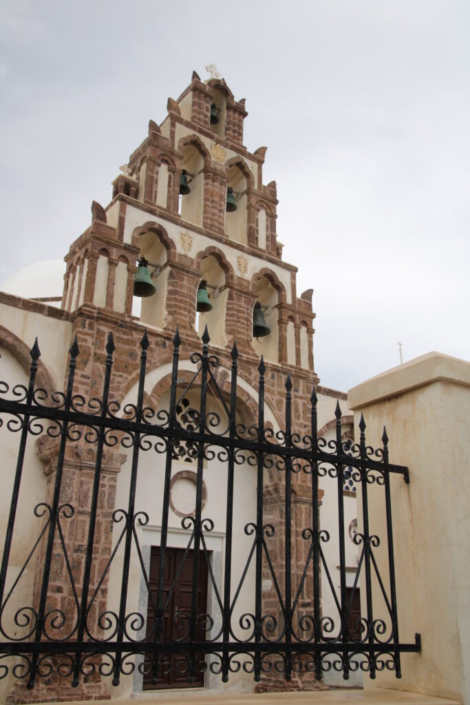 Ornate white stucco facade of the Church of Panagia Mesiana in Emporeio, fronted by a black wrought iron gate; the tall arched structure is framed with red stone columns and topped with three tiers of bells in a 3-2-1 arrangement, with a small wooden door set into the central archway
