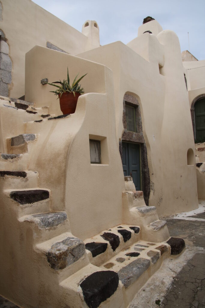 Curving view of residential structures within Emporeio Castle, with smooth white stucco walls, rounded facades, and sculptural rooftops at different levels; in the foreground, a steep, narrow stairway without a railing climbs between wooden doors and shutters, with a large spiky succulent in a red pot perched on a small landing
