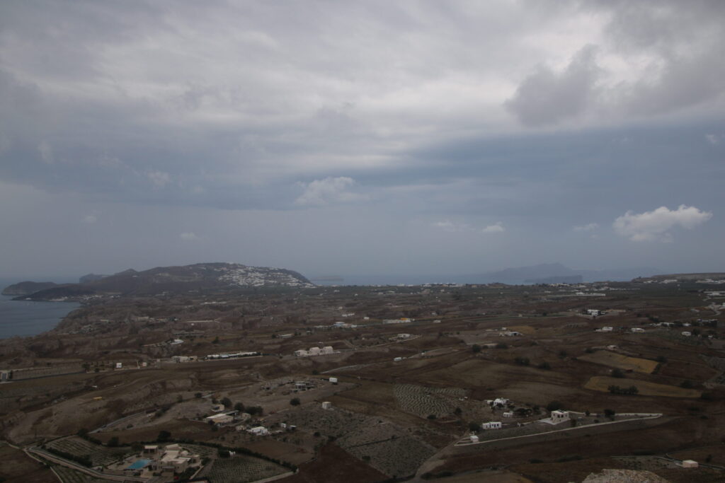 View from Prophet Elias showing a distant landscape of tiny white buildings tapering to a point on the left near the horizon, with a glimpse of the caldera below; the sky above is layered with shifting greys, with hints of blue and sunlight trying to break through