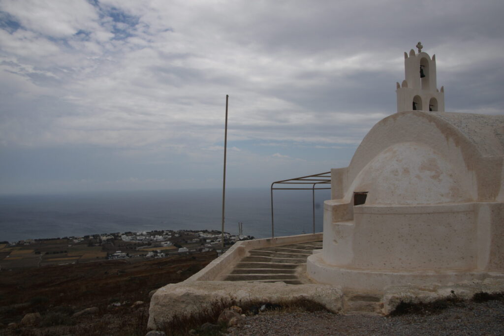 Mottled tan-and-white stucco chapel at Prophet Elias Hamilos with a sculptural, arch-shaped structure set on a square base, surrounded by a curved retaining wall; a bell tower with three bells in arched openings sits atop the chapel; behind it, the ground slopes away to distant, tiny buildings and a misty strip of sea beneath a textured grey sky and a few patches where the blue peeks through