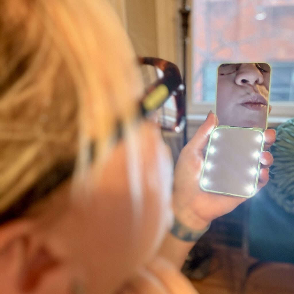 A sleek LED-lit compact travel mirror opened to show its dual sides — magnification on one, shown off with Ginny kissy face, standard reflection on the other.
