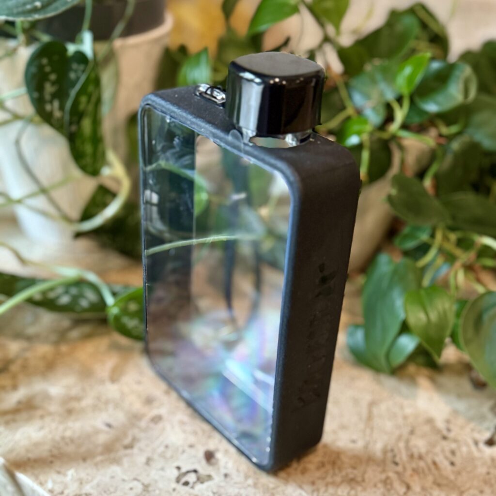 Book-shaped water bottle, clear with black edges, sitting in from of some leafy plants.