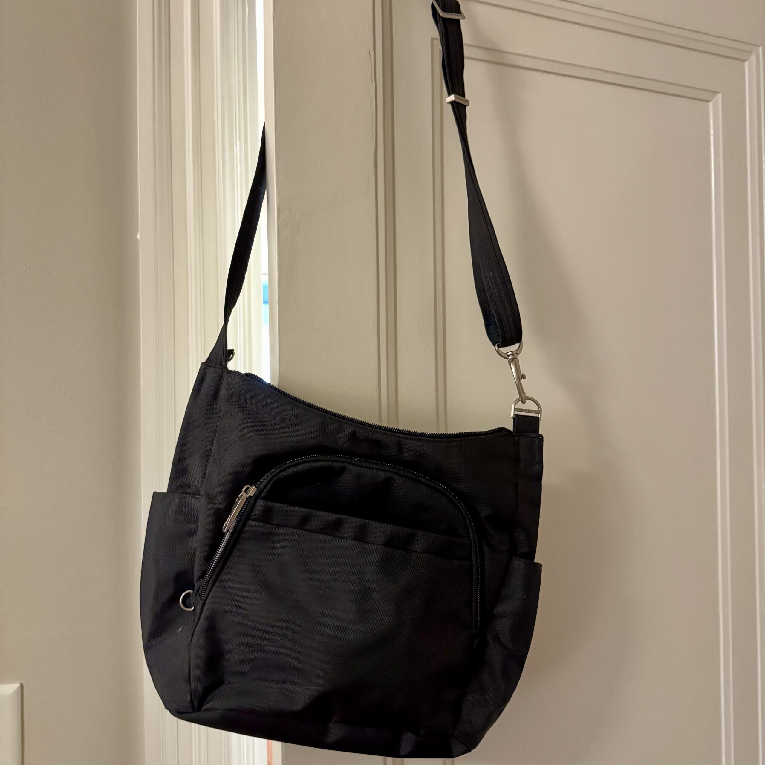 A black Travelon Anti-Theft Crossbody hangs on a door, showing its roomy size and ample exterior pockets.