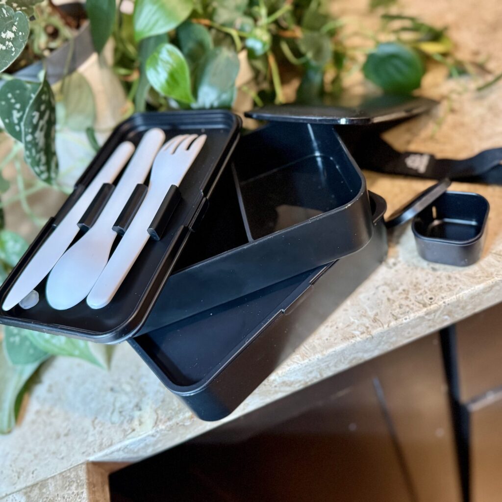 Three layer bento box, showing the airtight-lid on one layer, the dividers on the second, and the three pieces of silverware that clip neatly into the third, with the top lid, sauce container, and elastic strap to hold it all together.