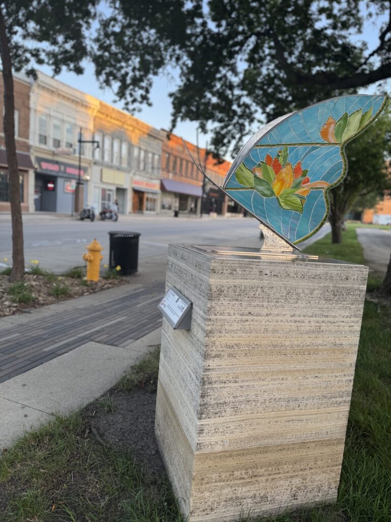 Colorful stained-glass mosaic wing sculpture on a limestone plinth along Main Street, Mason City.