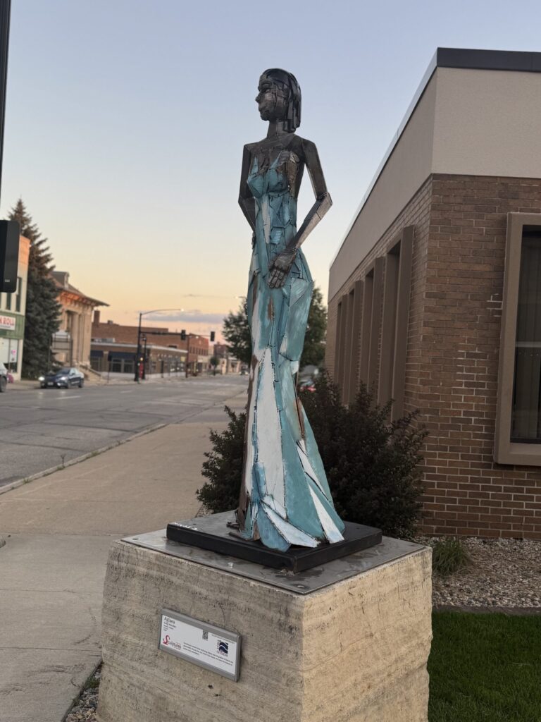 Metal sculpture of a woman in a turquoise dress on a downtown Mason City sidewalk at dusk.