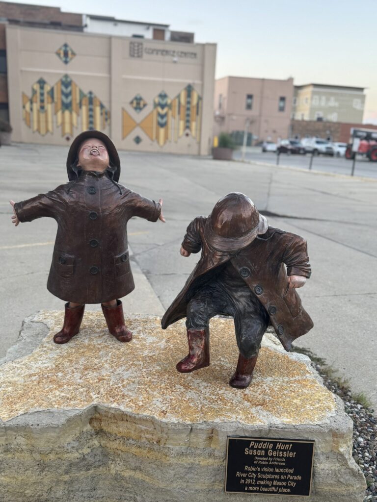 Bronze “Puddle Hunt” statues of two children in raincoats with a geometric mural in Mason City.