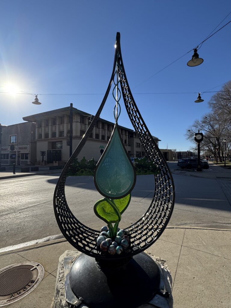 Glass-and-metal teardrop sculpture on a downtown corner with the Historic Park Inn behind, Mason City, Iowa.